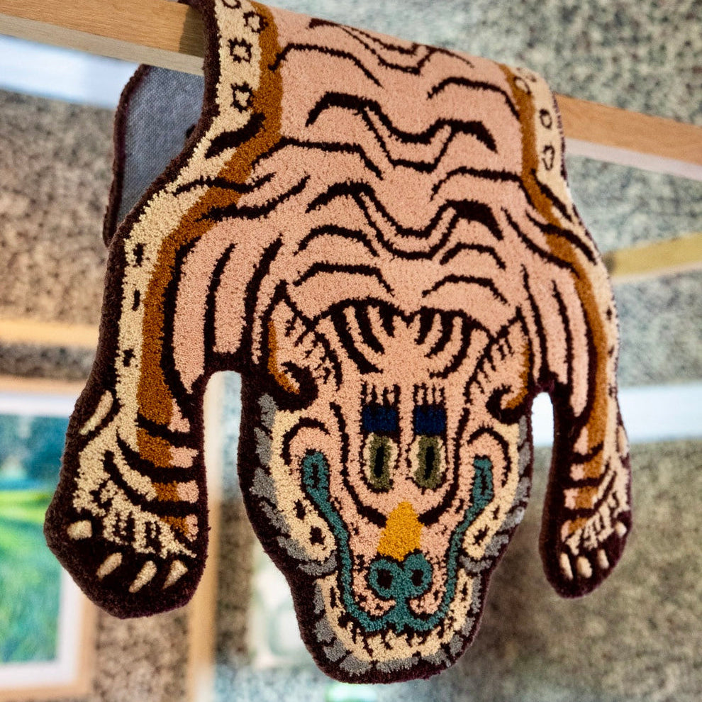 A hand-knotted wool textile piece shaped like a tiger's head, The design features pink and brown stripes with teal and gold accents on the face.