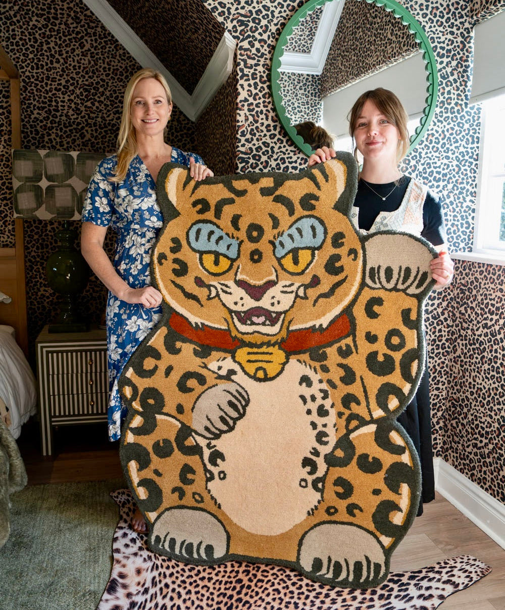 A large, maneki neko-style leopard-shaped rug or mat is held up by two women in a room with matching leopard-print wallpaper. The rug features a tan and beige base with black leopard spots, blue eyes, and a red collar.