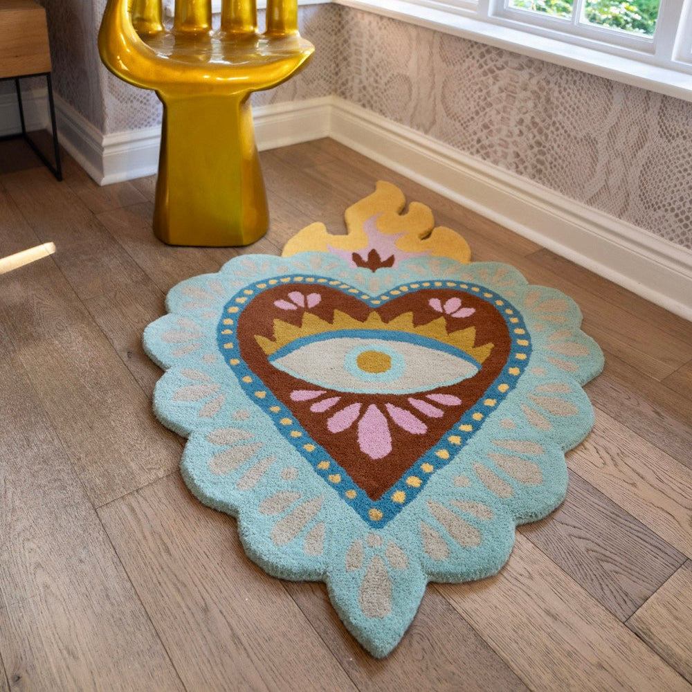A heart-shaped area rug with a central eye motif and scalloped edges, styled on a wooden floor in a bright room next to a gold hand-shaped chair.