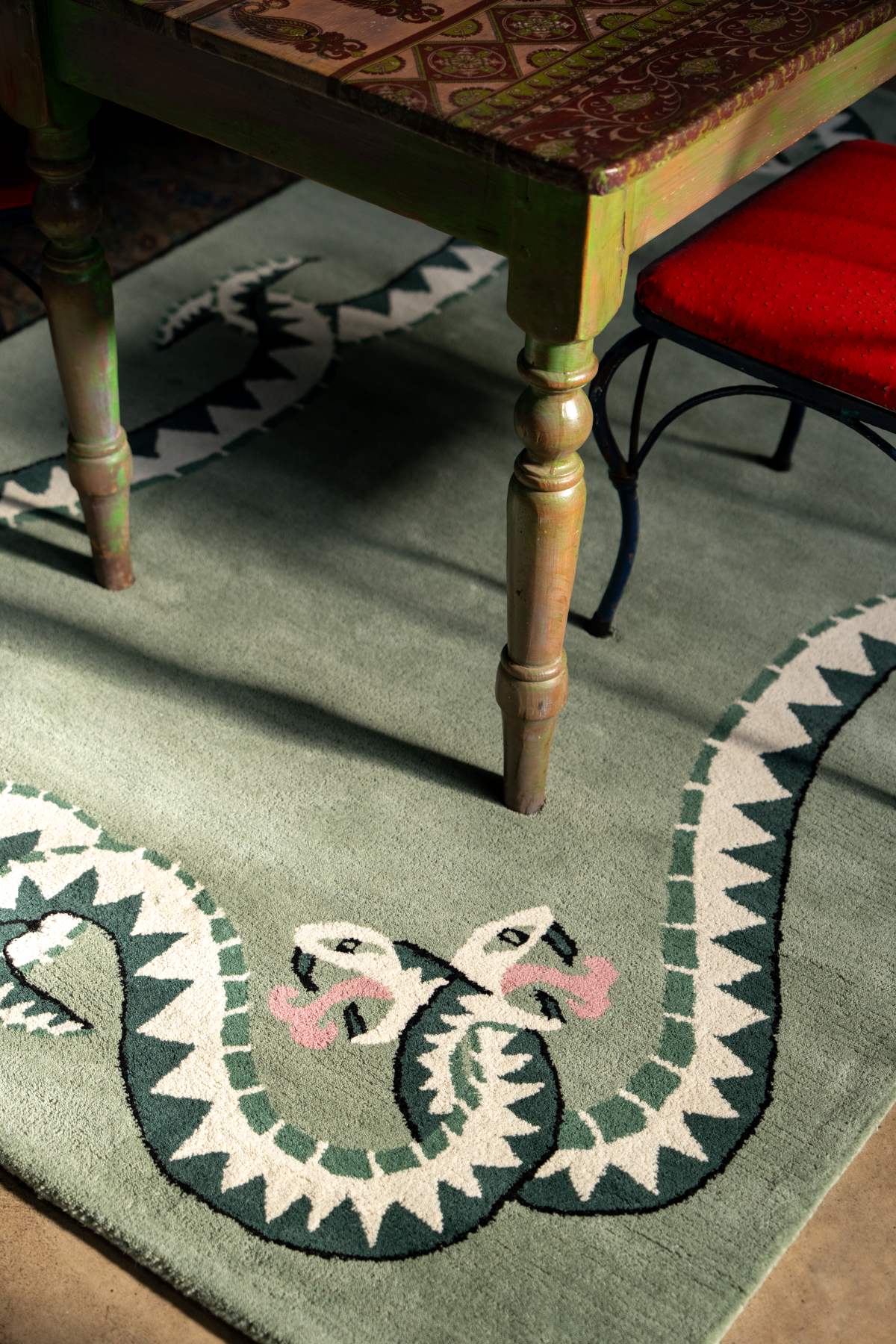Decorative green rug with snake design on the floor, table with patterned top in the background.