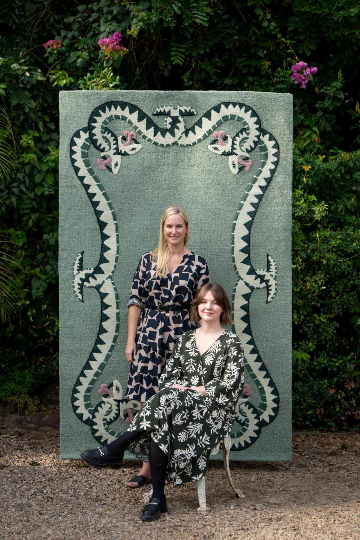 Two south african women in patterned dresses standing in front of a large decorative green rug with snake designs.