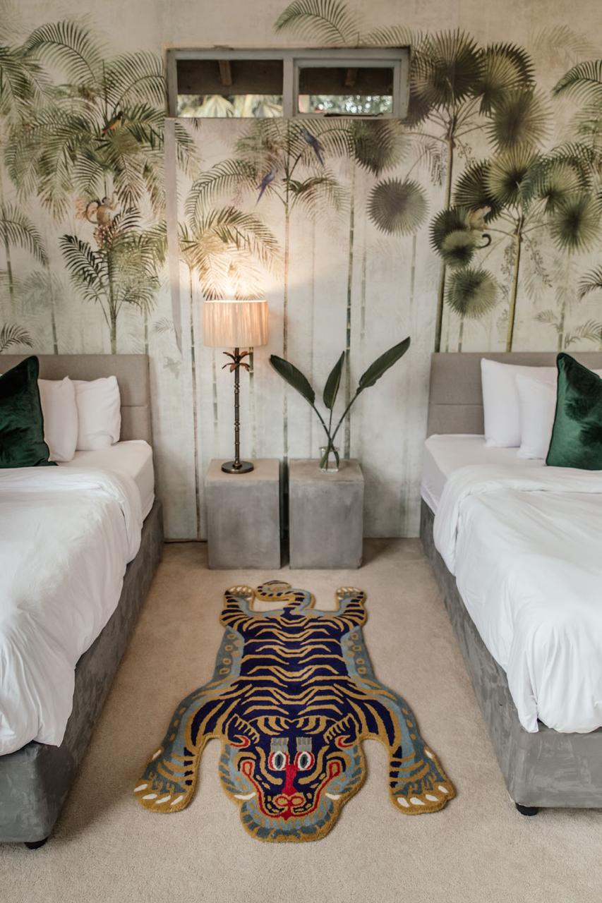 Tropical-themed bedroom with palm tree wallpaper, two beds, a lamp, and a colorful tiger wool rug.